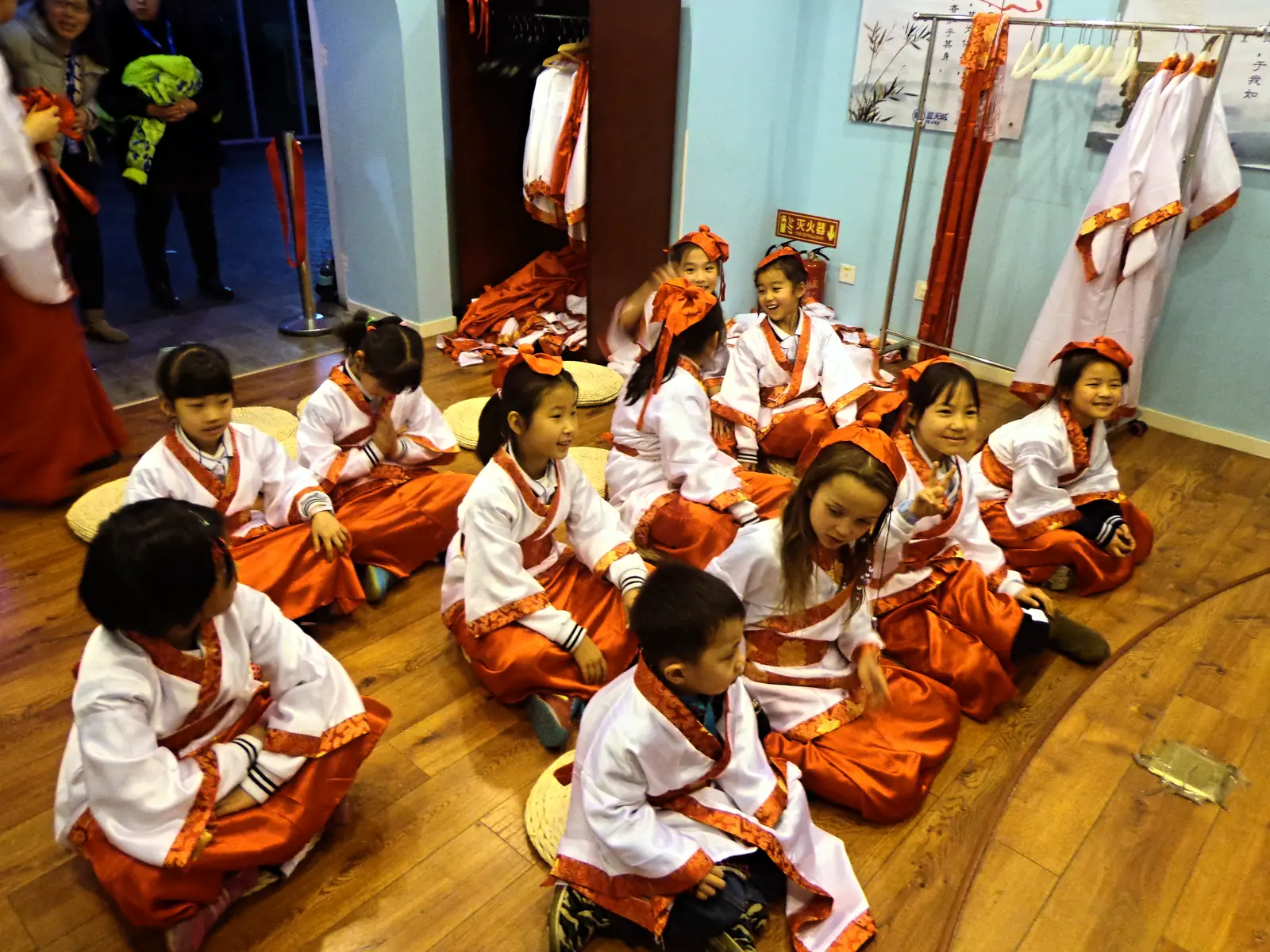 Children in orange and white hanfu seated on floor cushions for a traditional dress activity