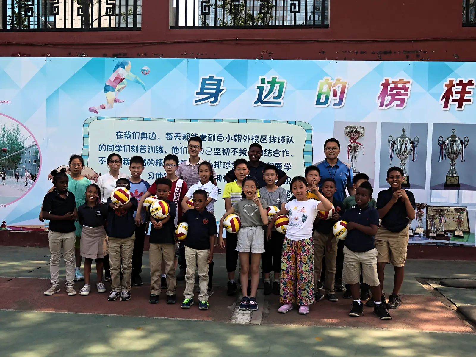 Pupils holding volleyballs at a school assembly, trophies displayed behind them